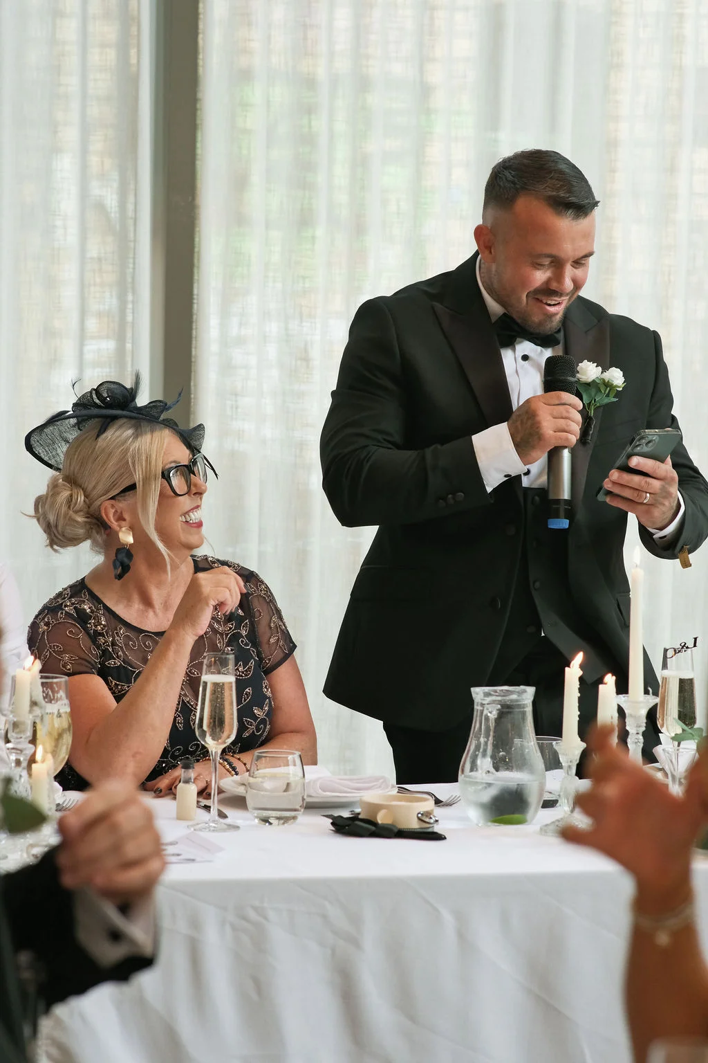 A groom in a black tuxedo delivers a speech holding a microphone and mobile phone, while a guest wearing a dark fascinator looks on and laughs at the wedding reception table.