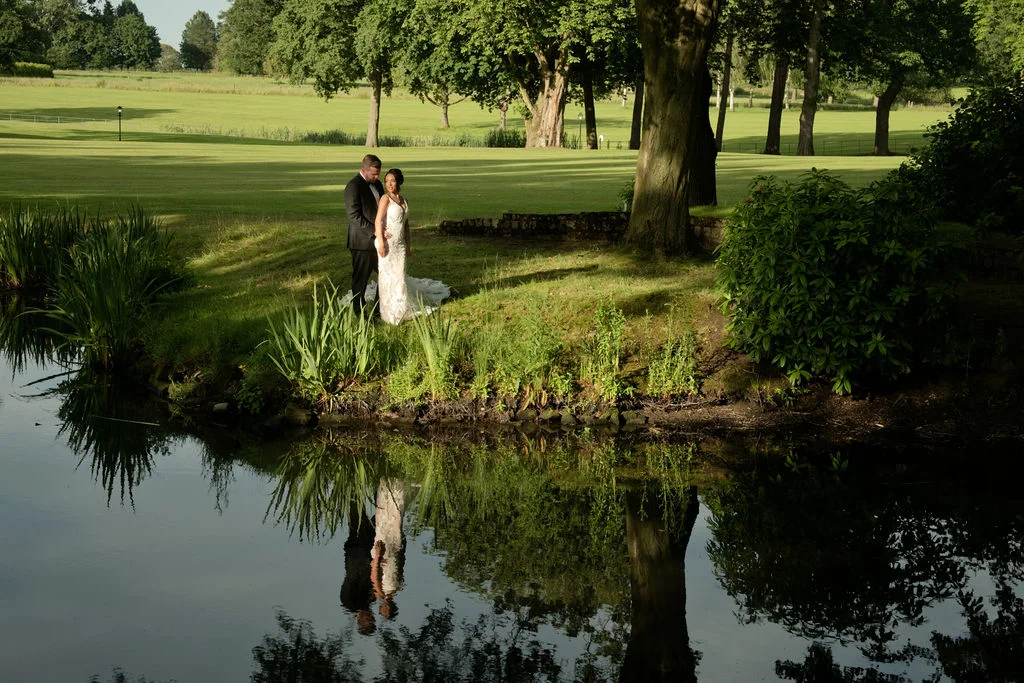 A newlywed couple stands together by a serene lakeside on the grounds of a country estate, captured in golden hour light with a striking reflection in the water.