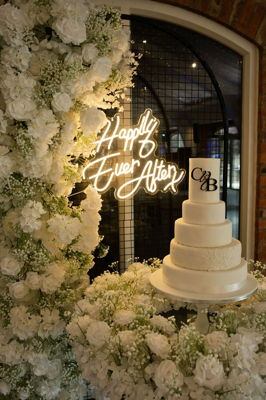 A stunning multi-tiered white wedding cake rests on a table surrounded by lush white roses and baby's breath, positioned in front of a glowing neon sign and a dramatic floral wall.