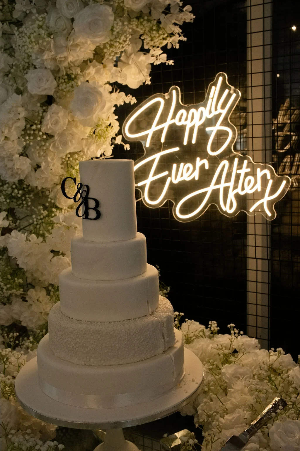 A stunning five-tier all-white wedding cake sits before a lush white floral arch and a glowing neon sign reading Happily Ever After.