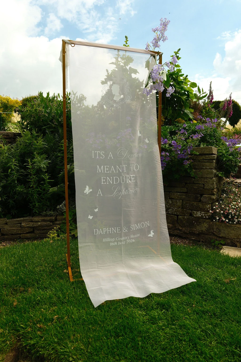 A customized sheer fabric welcome sign hangs from a copper frame outdoors in a lush garden setting against a dry stone wall.