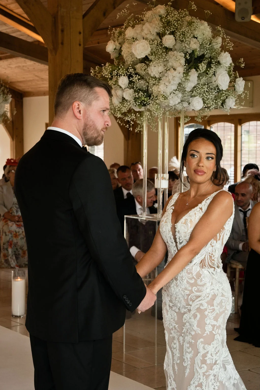 A bride and groom holding hands during their civil ceremony inside an oak framed barn, featuring a large white floral arrangement overhead.