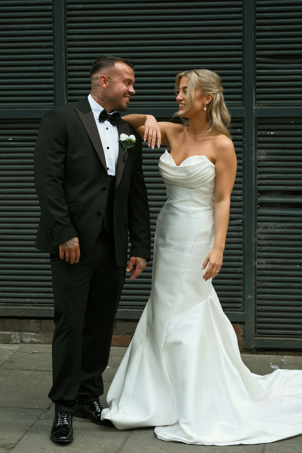 A stylish newlywed couple shares a joyful glance outdoors against a dark slated backdrop, with the groom in a classic black tuxedo and the bride wearing an elegant strapless satin gown.