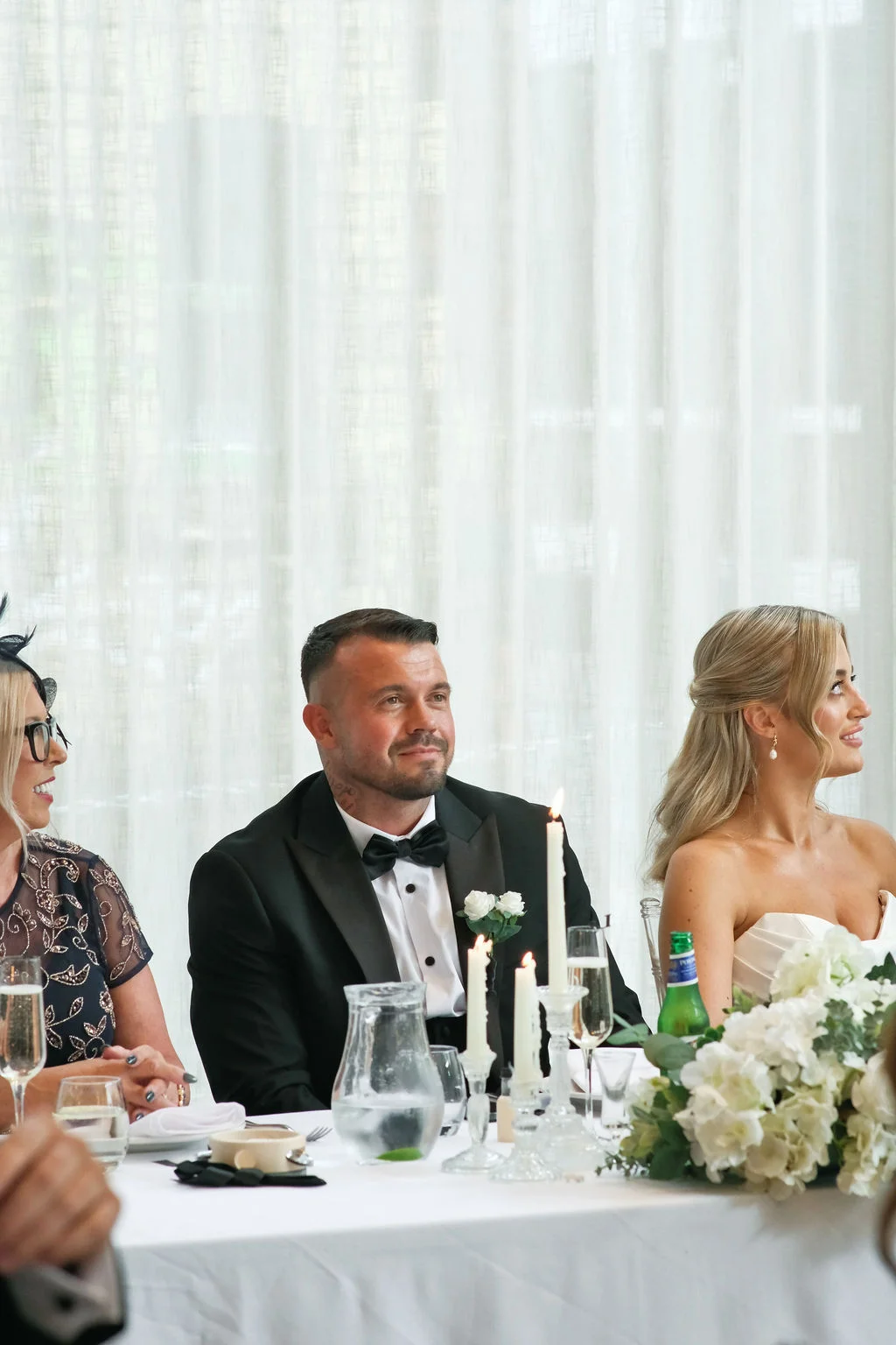 Newlyweds and a guest seated together at a beautifully set wedding reception top table featuring white floral arrangements and glowing taper candles.