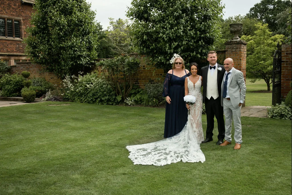 A bridal party and guests pose for a formal portrait on a manicured lawn outside a historic country house with a stone garden wall.