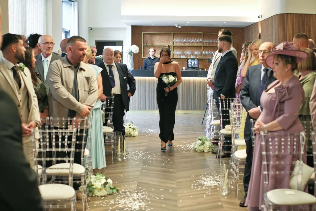 An emotional bride in a black strapless column dress walks down the aisle holding a bouquet of white roses while guests look on from transparent chairs.