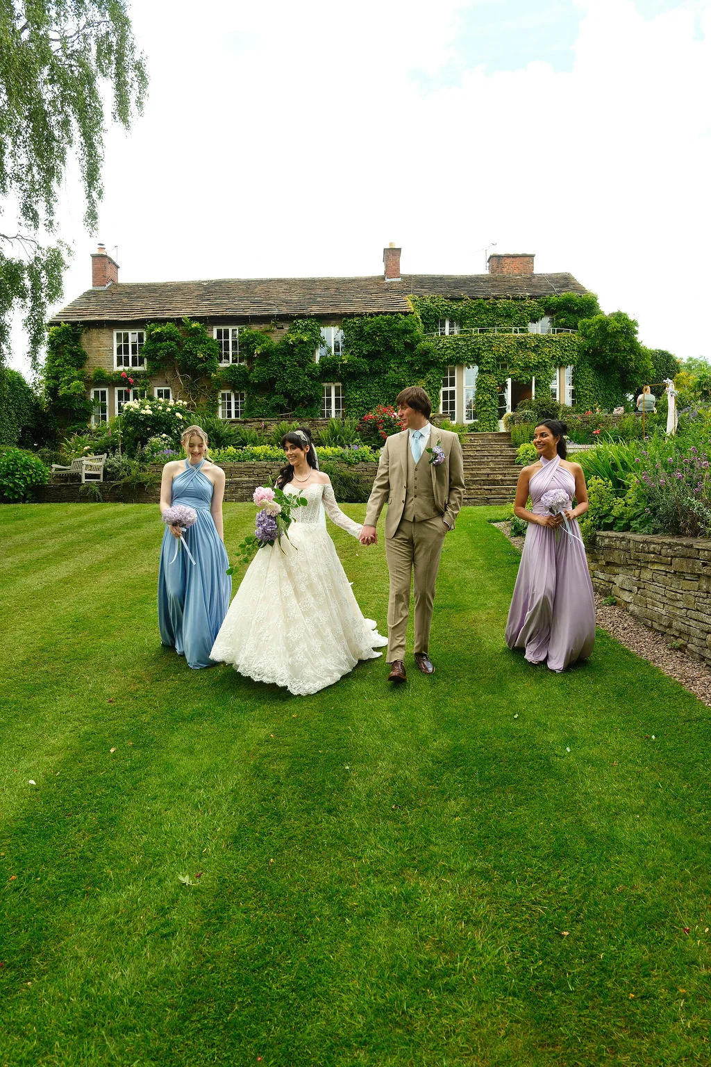 A bride in an off-the-shoulder lace gown and groom in a beige suit walk hand in hand across a manicured lawn, flanked by bridesmaids in blue and lilac dresses in front of an ivy-covered country house.