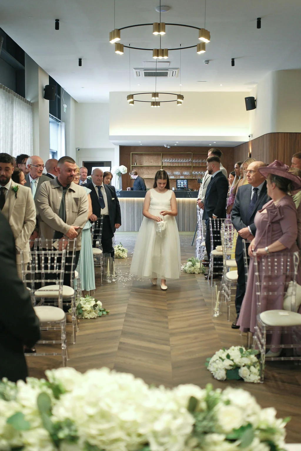 A young flower girl walks down the aisle carrying a small basket between rows of seated guests in a modern hotel wedding venue.