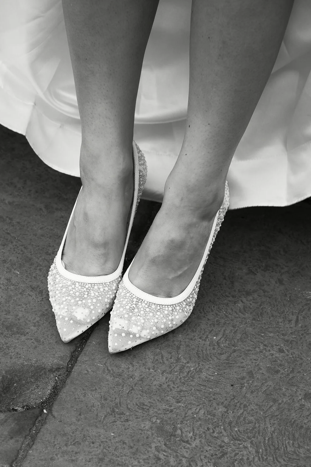 A close-up black and white photograph showcases a bride's feet wearing intricately embellished pointed-toe wedding shoes against a stone floor.
