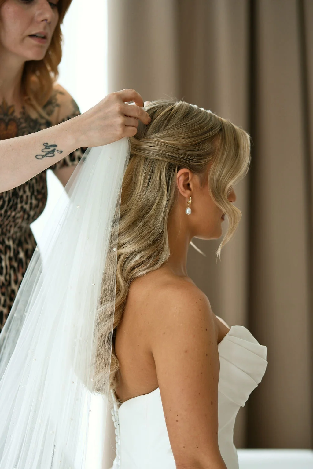A bride with a half-up hairstyle and soft waves has a pearl-embellished veil carefully placed in her hair during wedding morning preparations.