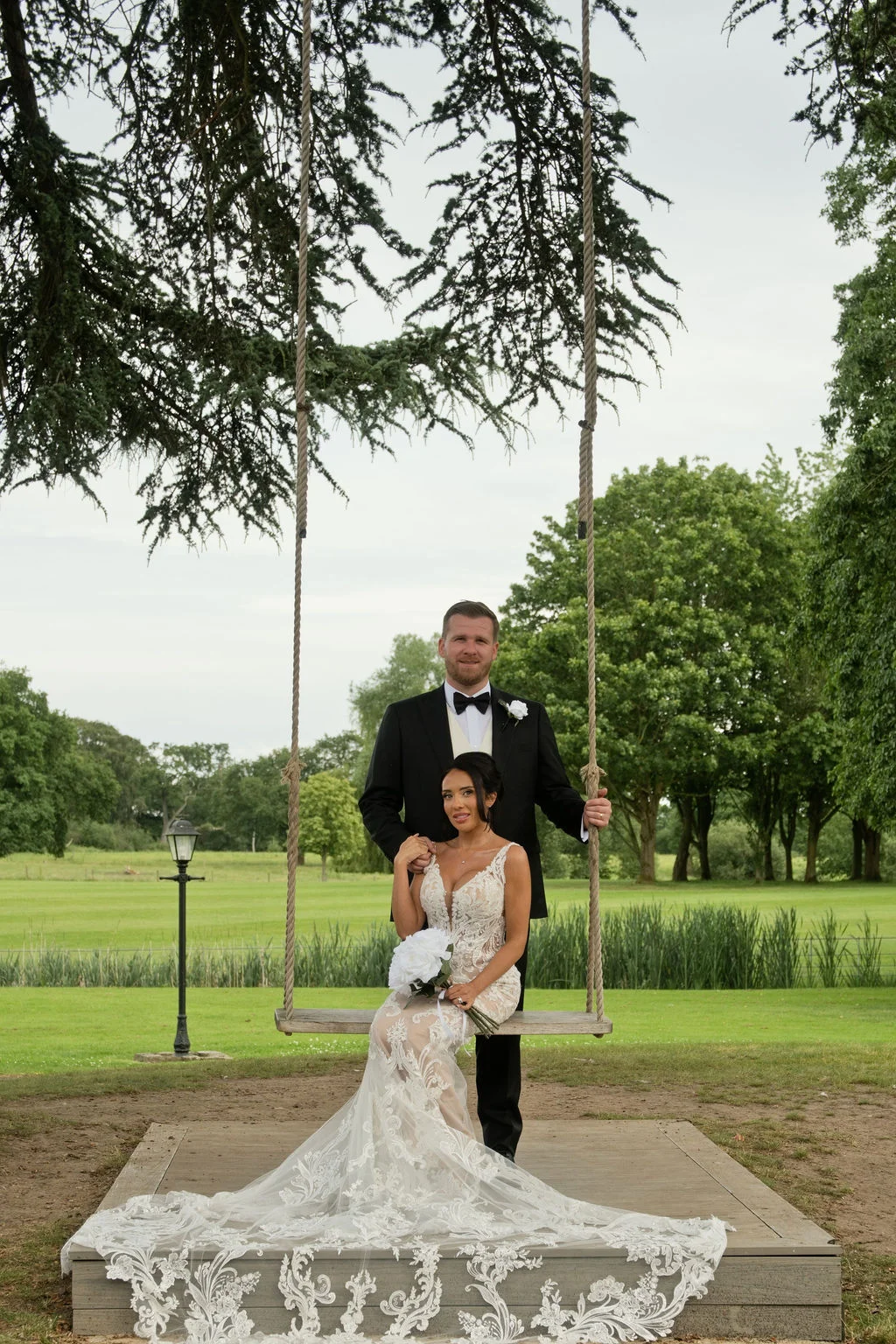 A newlywed couple poses together outdoors on a rustic wooden rope swing beneath a large leafy tree, with the bride seated in an intricate lace gown and holding a white bouquet while the groom stands behind her in a classic black tuxedo.