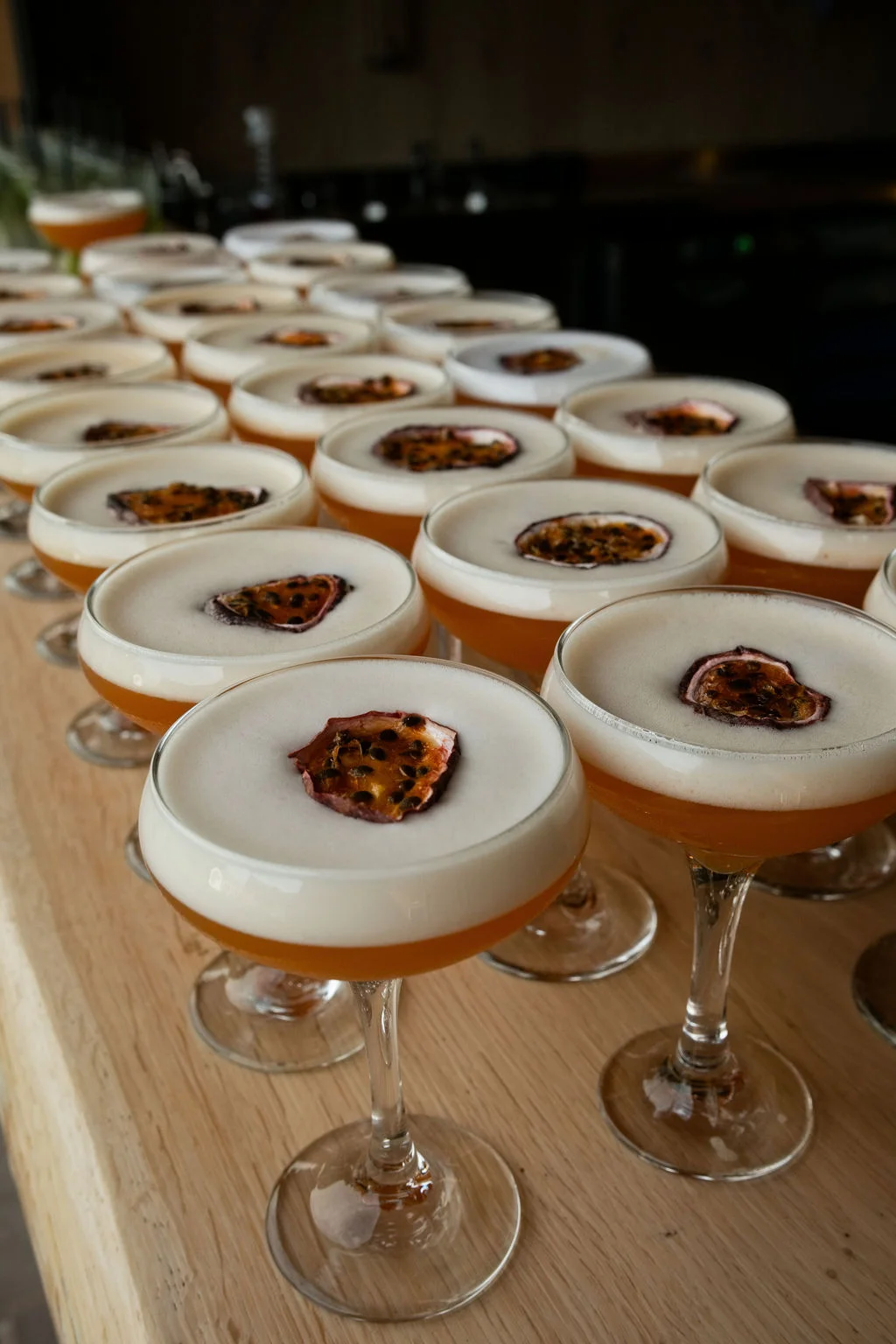 An array of freshly prepared pornstar martini cocktails garnished with dehydrated passion fruit slices sits lined up on a wooden bar during a wedding drinks reception.
