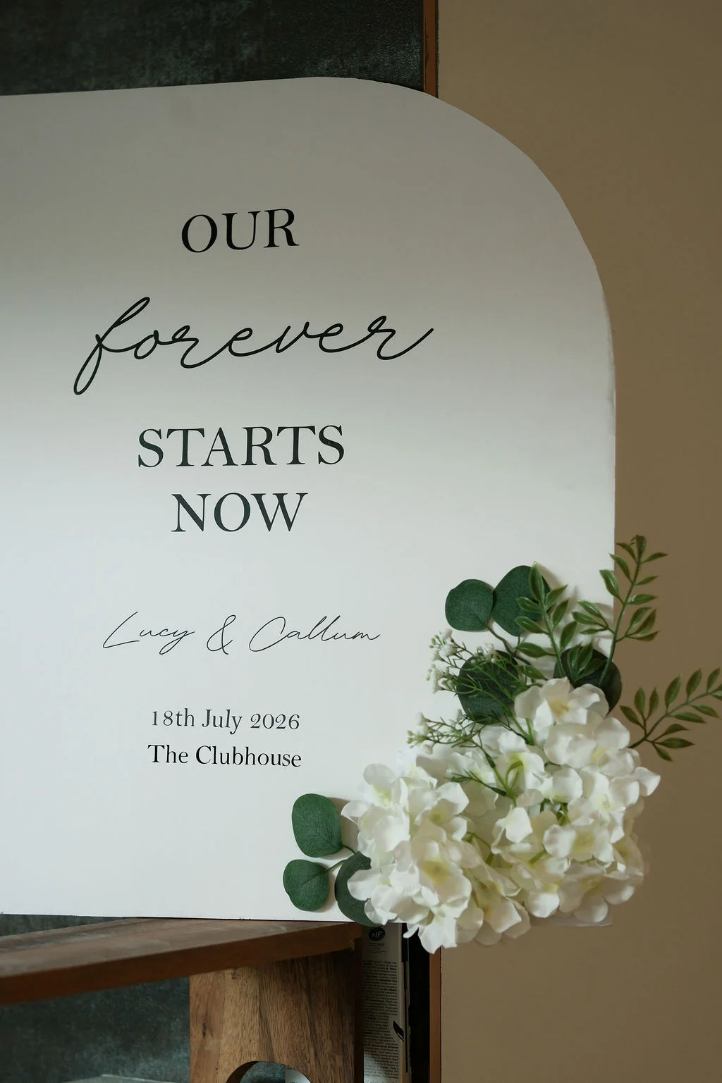 An arched wedding welcome sign featuring black typography and white floral arrangements with eucalyptus foliage sits on a wooden stand.