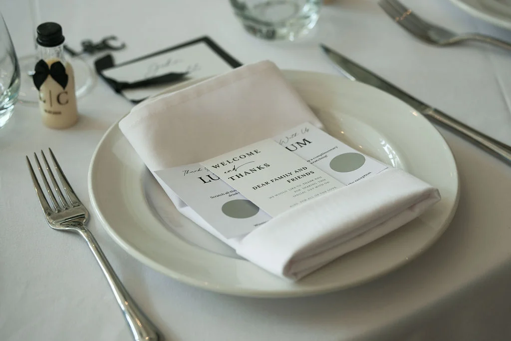 An elegant wedding place setting featuring a crisp white plate, neatly folded napkin, stationery cards, and a miniature favour bottle with a black bow.