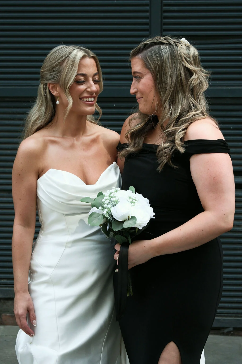 Two brides share a joyful moment together outside, with one wearing a white satin strapless gown and the other in a black off-the-shoulder dress holding a white rose bouquet.