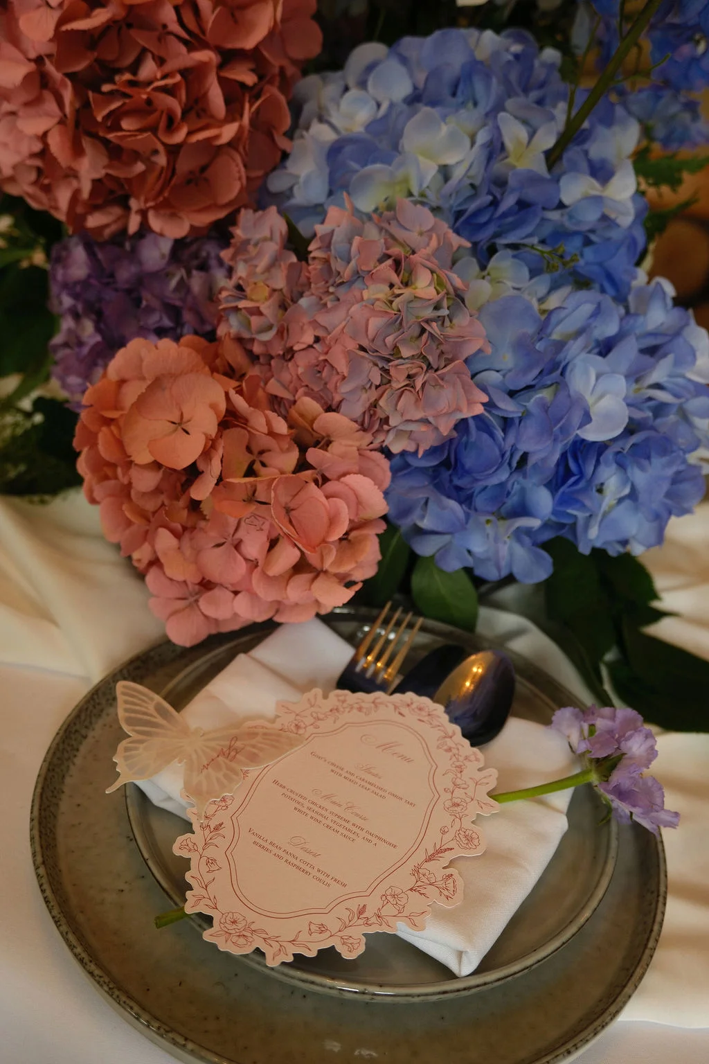 An elegantly styled wedding place setting features a custom illustrated menu card adorned with a delicate butterfly motif, placed atop layered plates with lush hydrangea floral arrangements in pink and blue tones.