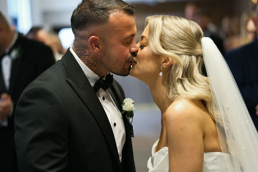 The newly married couple share their first kiss, with the groom wearing a classic black tuxedo and the bride in a strapless gown with pearl-embellished veil and half-up hair.