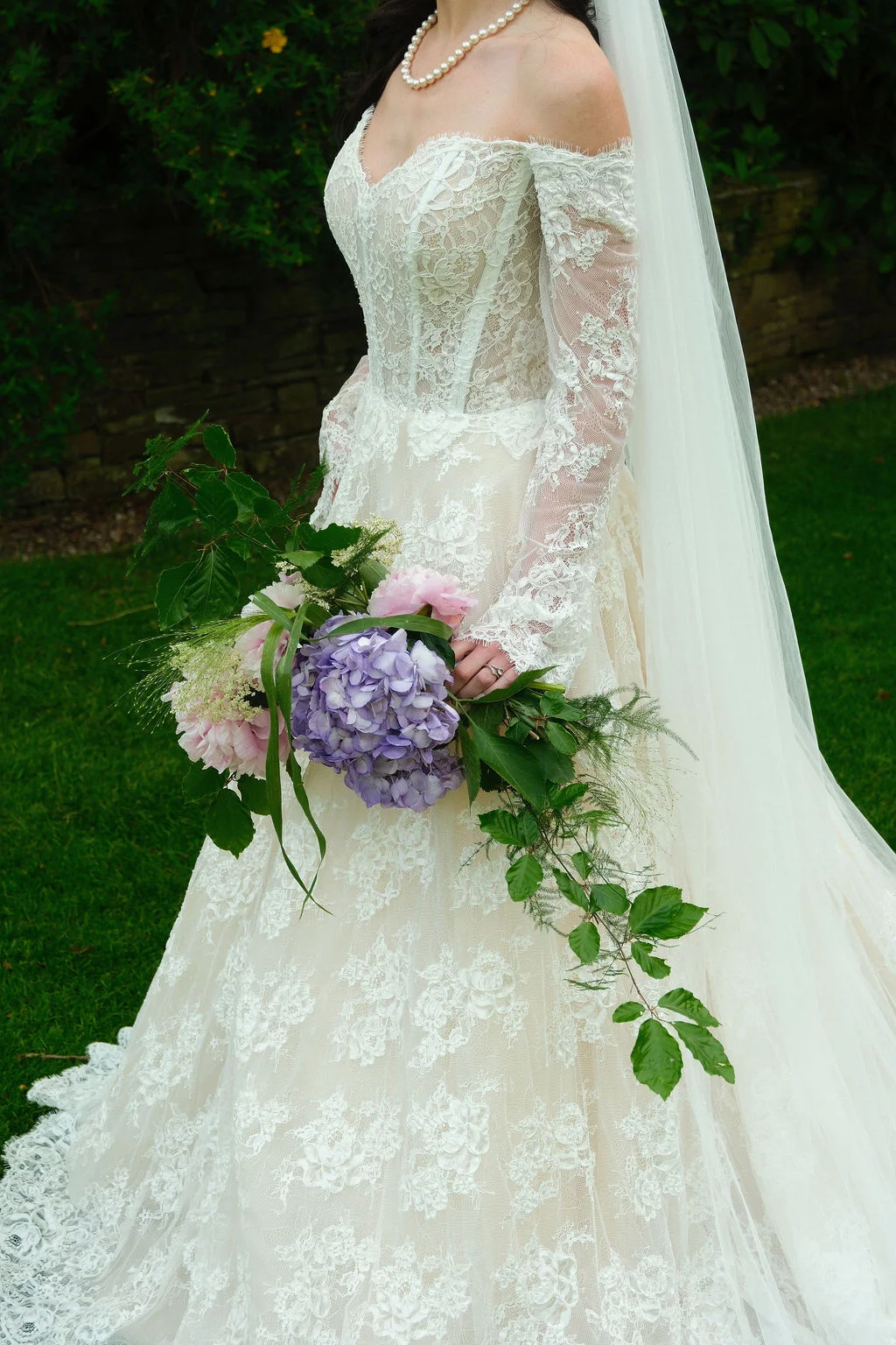 A bride in an off-the-shoulder lace gown with long sleeves holds a trailing cascade bouquet featuring purple hydrangeas and green foliage outdoors.
