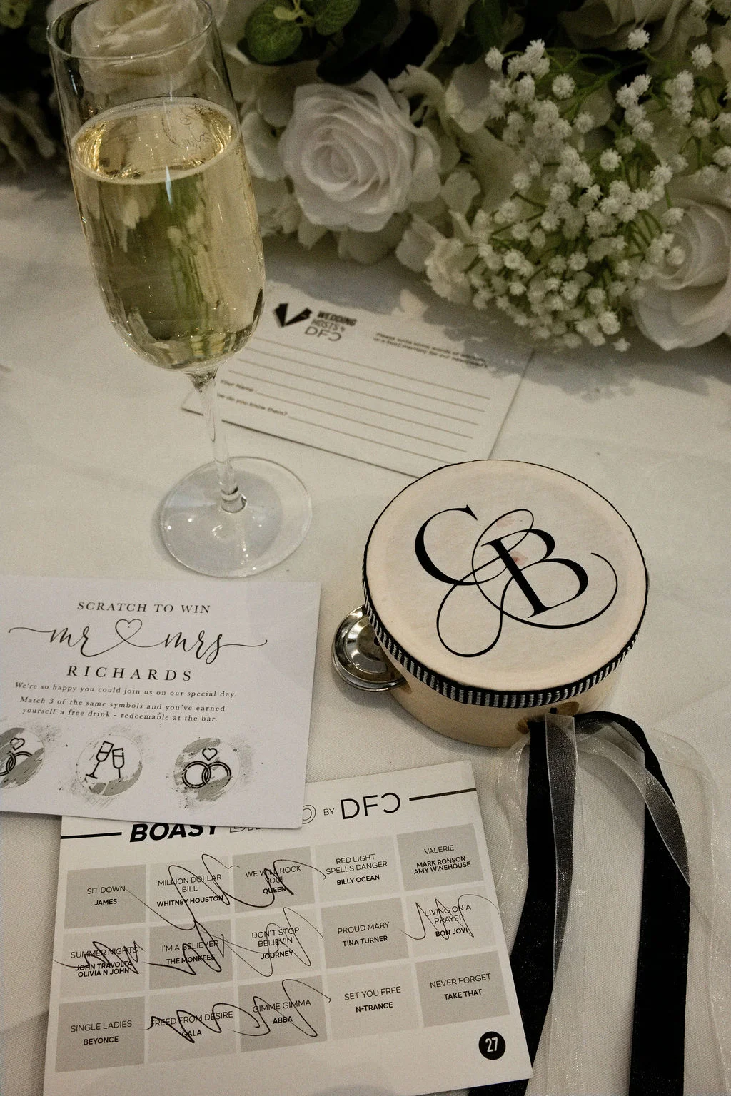 An overhead flat lay of a wedding reception table featuring a flute of champagne, personalised stationery, a branded tambourine with a monogram, and white floral arrangements.