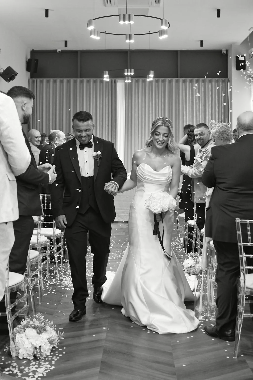 A newlywed couple joyfully walks back down the aisle together hand in hand while guests shower them with confetti in a bright venue.