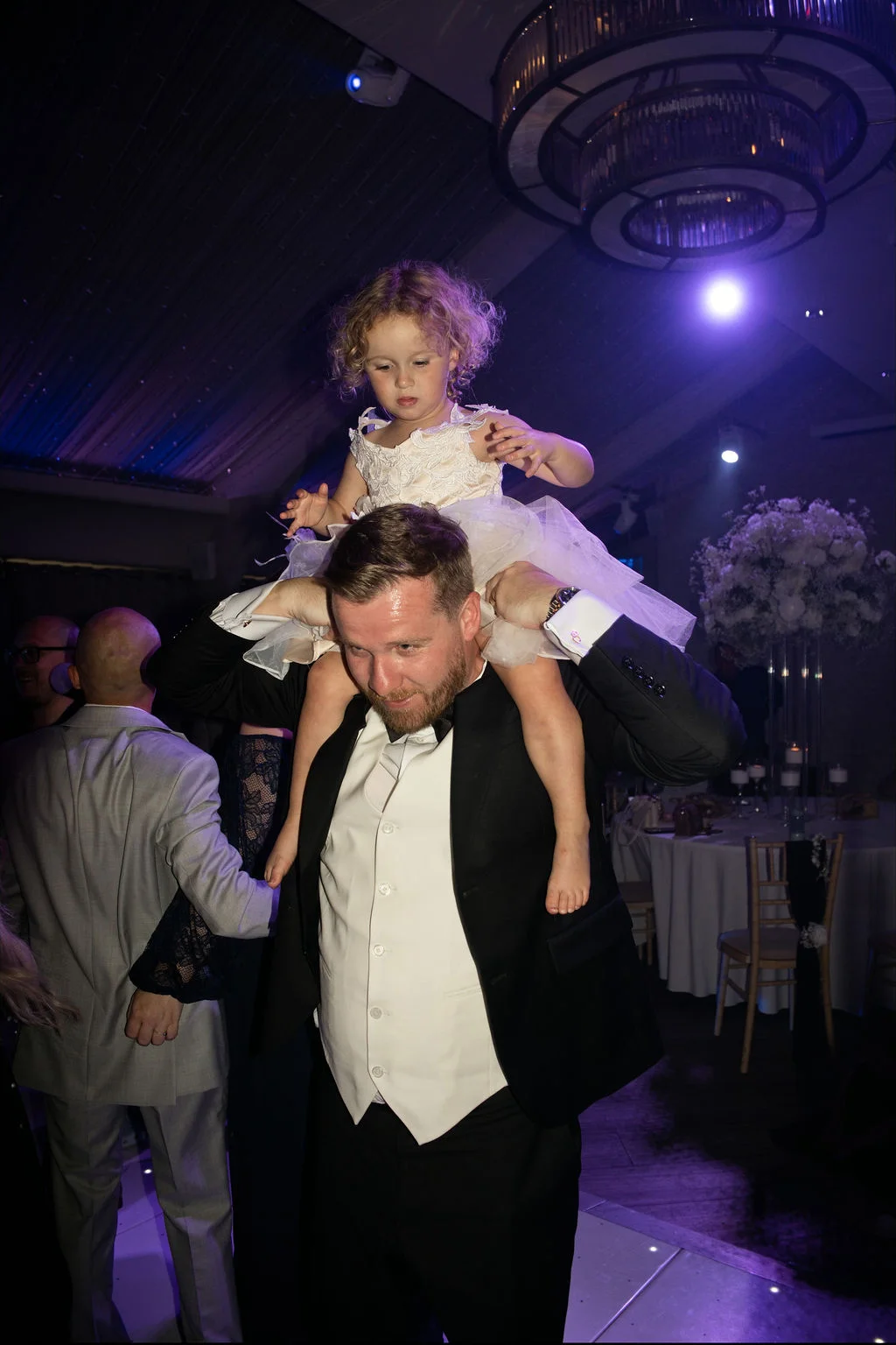 A young flower girl in a white tulle dress enjoys a shoulder ride from a guest wearing a tuxedo on the dance floor during an evening wedding reception.