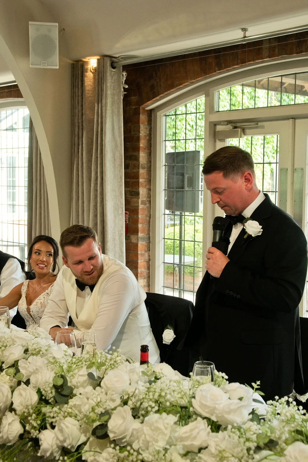 A groom delivers a speech into a microphone at the top table during a wedding reception, while the bride and groom look on and smile amidst a lush white floral runner.