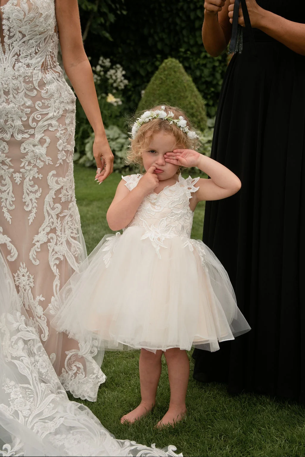A barefoot flower girl wearing a tulle dress and flower crown rubs her sleepy eyes while standing outdoors on the grass between a bride and a bridesmaid.