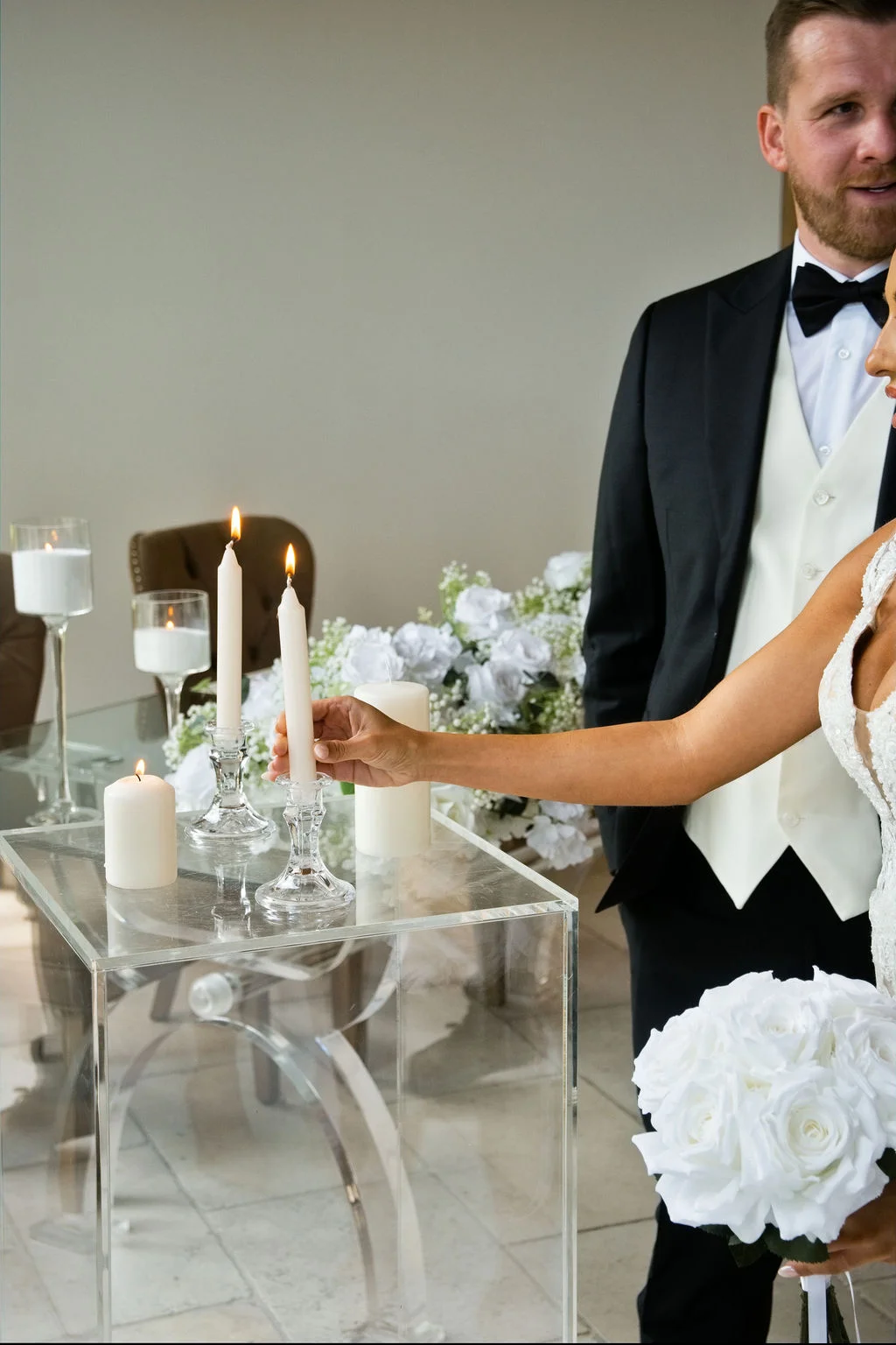 A bride and groom participate in a unity candle ceremony, with the bride lighting a taper candle on a modern clear acrylic stand next to white floral arrangements.