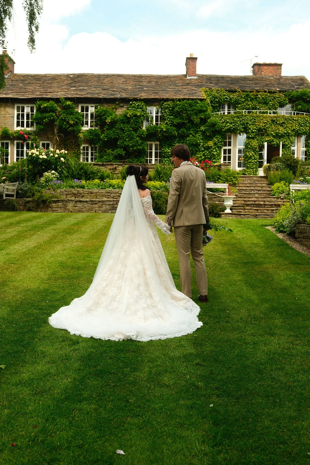 A bride and groom walk hand in hand across a manicured lawn toward a historic stone country house draped in lush green ivy.