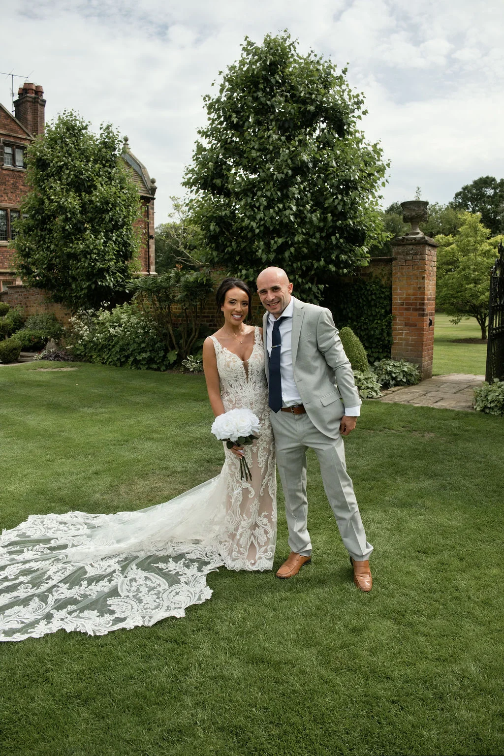 A smiling bride and groom pose together on a manicured lawn outside a historic country house, showcasing the bride's dramatic lace gown and long train.