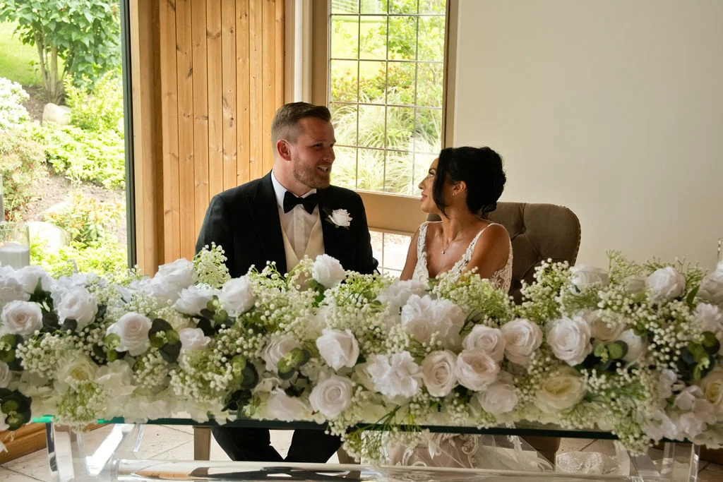 A smiling bride and groom share a tender glance across a lush white floral arrangement at their top table.
