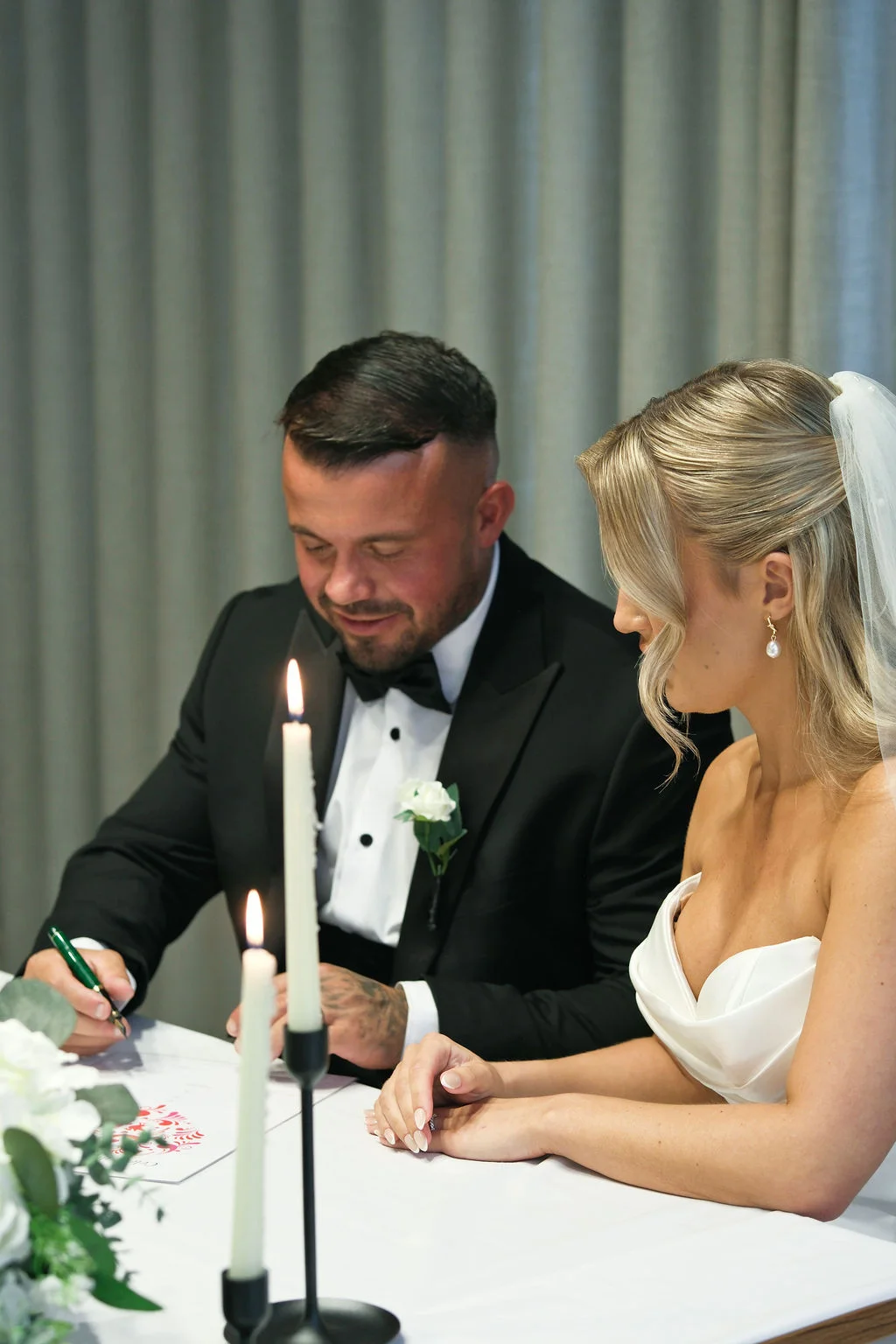 A newlywed bride and groom sit together at a table while signing the wedding register, illuminated by the soft glow of taper candles.