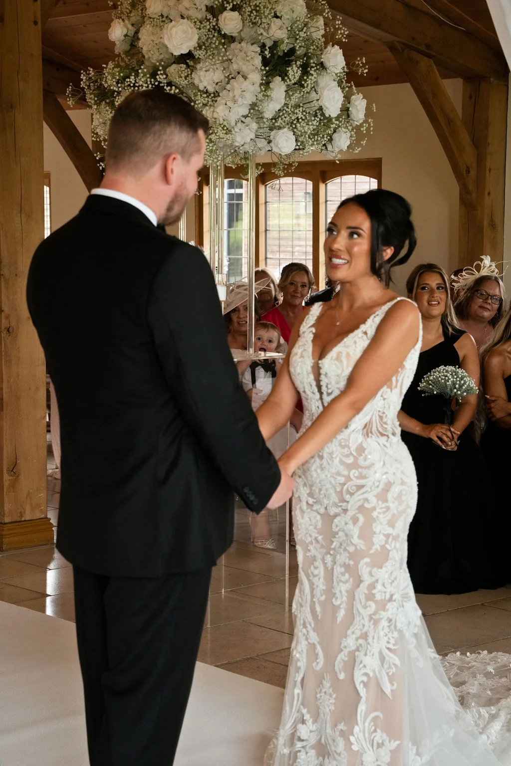 A bride in a fitted lace gown and groom in a black tuxedo hold hands and smile at each other during their indoor barn ceremony beneath a floral hanging installation.