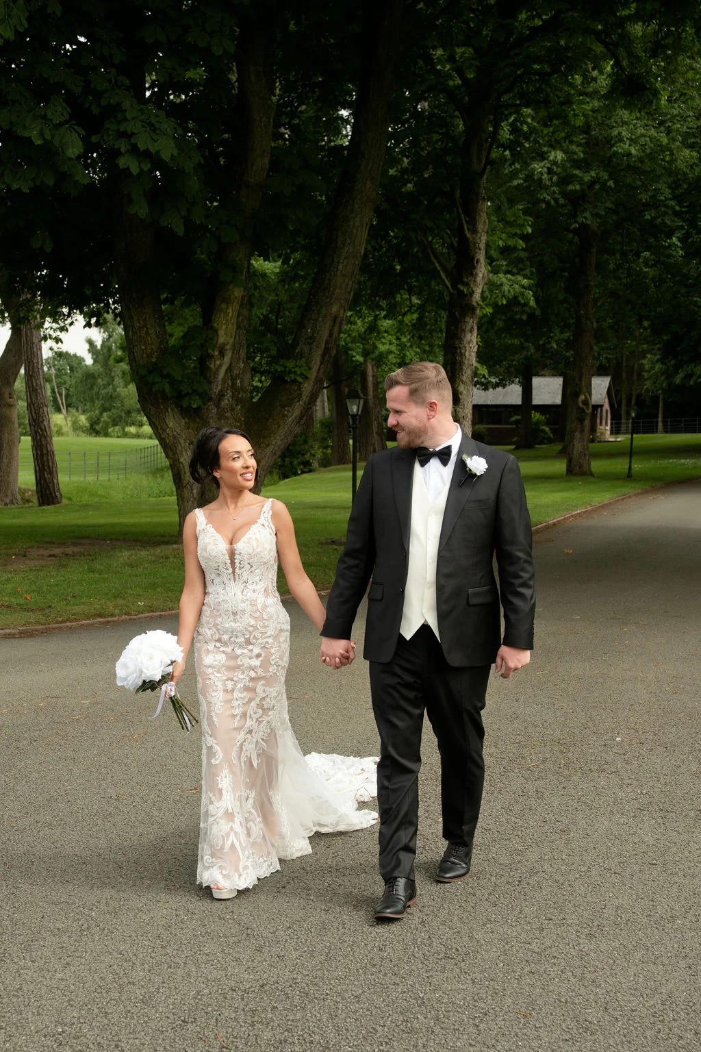 Newlyweds lovingly gaze at each other while walking hand-in-hand down a scenic tree-lined path.