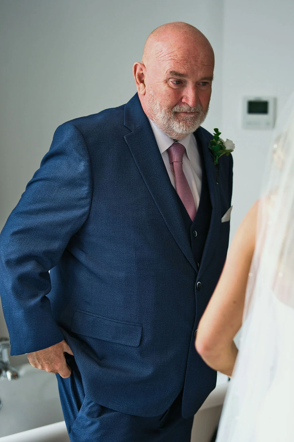 A mature father of the bride wearing a smart navy three piece suit and pink tie looks lovingly at his daughter in her wedding dress.