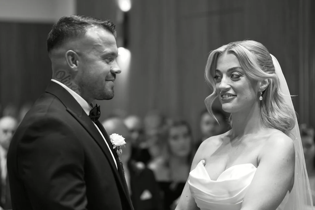 A black and white photograph captures the bride and groom sharing a tender glance during their wedding ceremony.