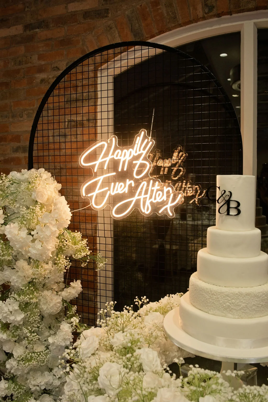 A multi-tiered white wedding cake sits beside a circular metal grid backdrop featuring a glowing neon sign and abundant white floral arrangements.
