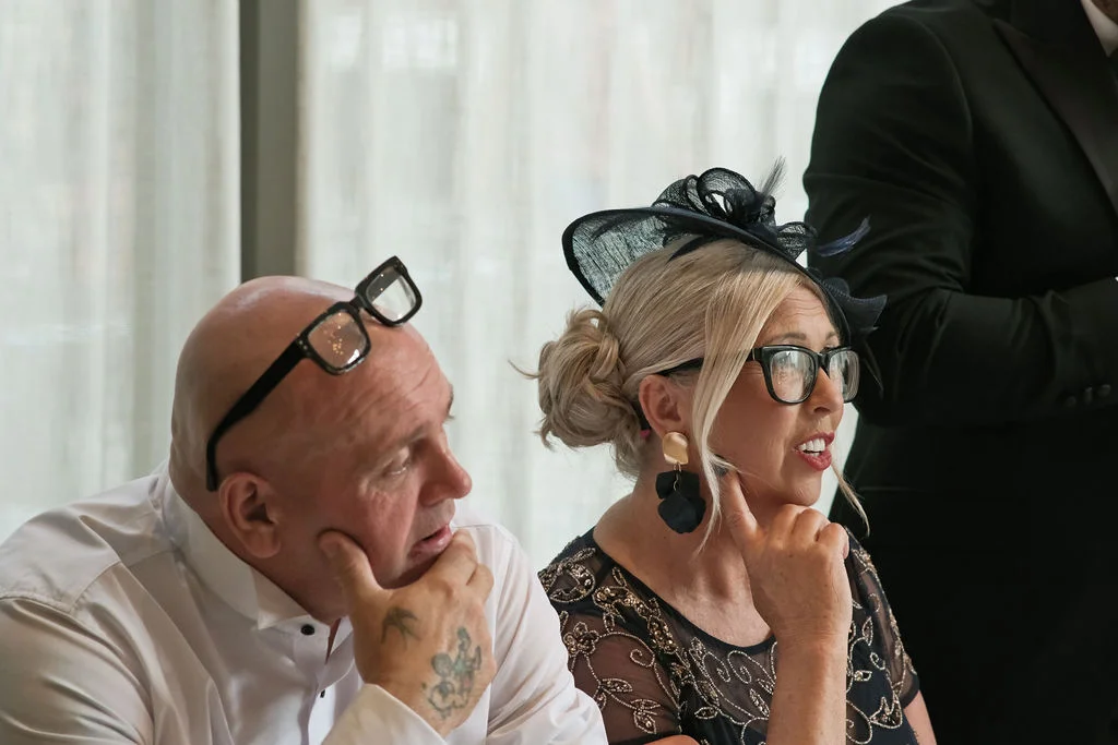 An older couple sitting together at a wedding reception, both wearing glasses and smiling while listening to speeches.