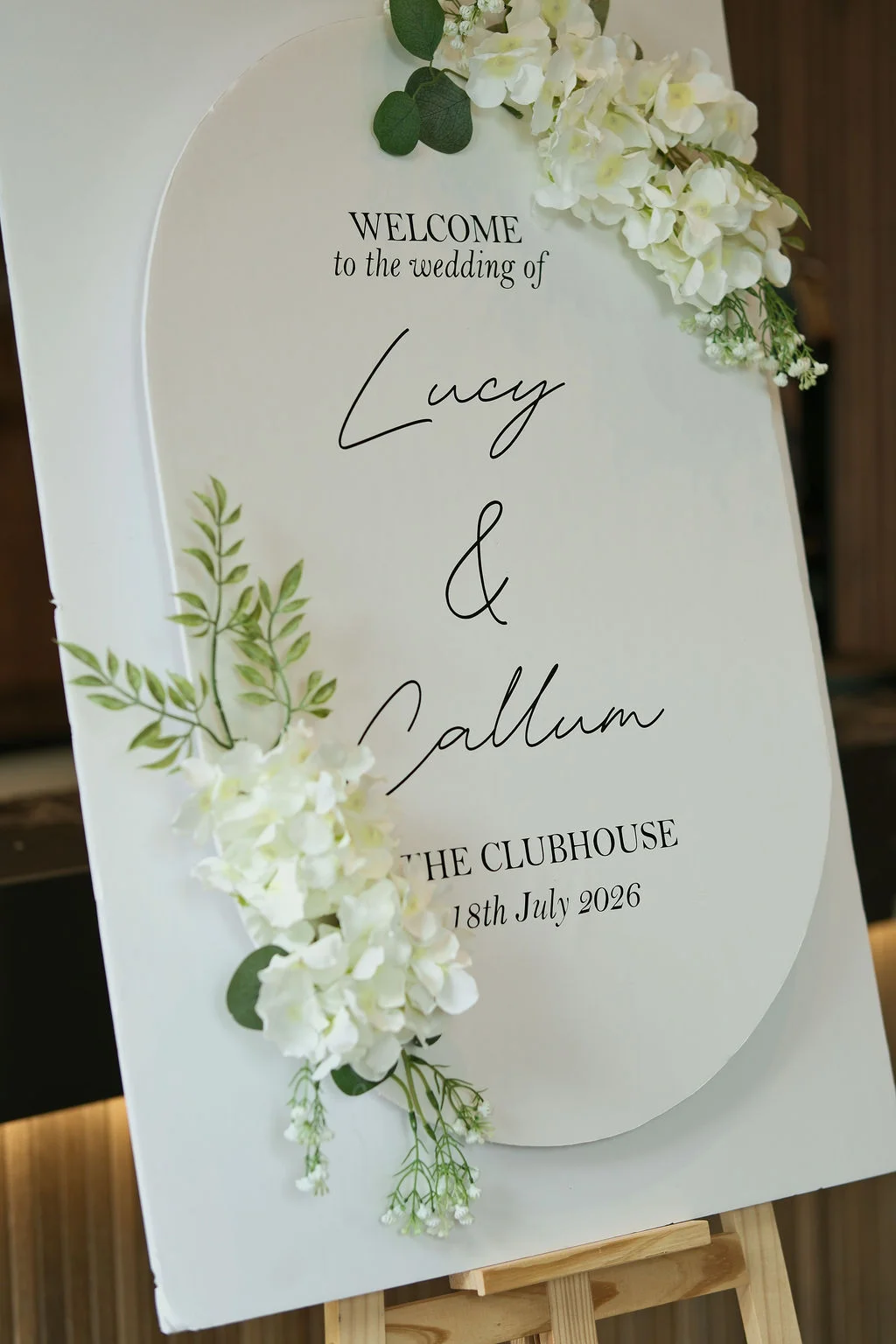 A modern arched white wedding welcome sign adorned with delicate white hydrangeas and green foliage stands proudly on a wooden easel.