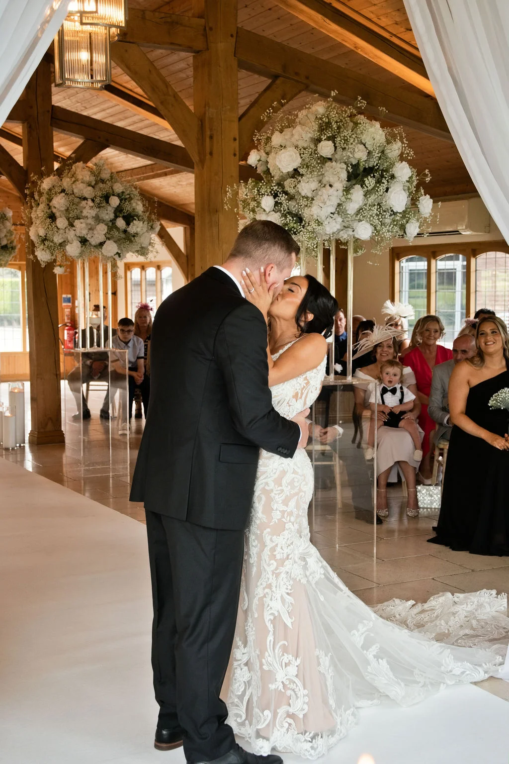 The newly married couple share their first kiss at the altar inside an oak framed barn venue decorated with white floral pedestal arrangements and ceiling drapes.