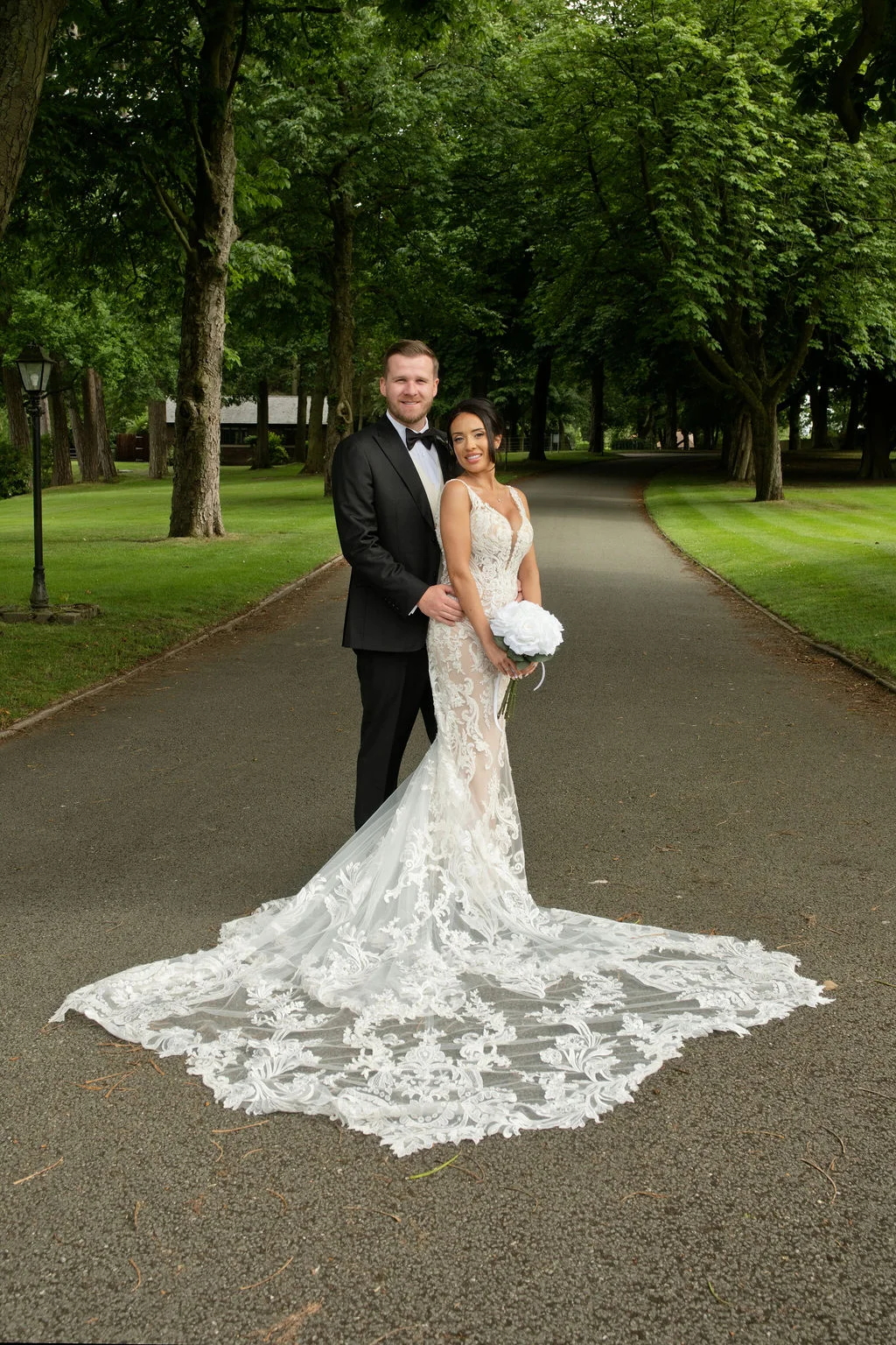A newlywed couple poses on a tree-lined driveway, with the bride wearing a stunning mermaid-style lace wedding dress featuring a dramatic, intricate train.