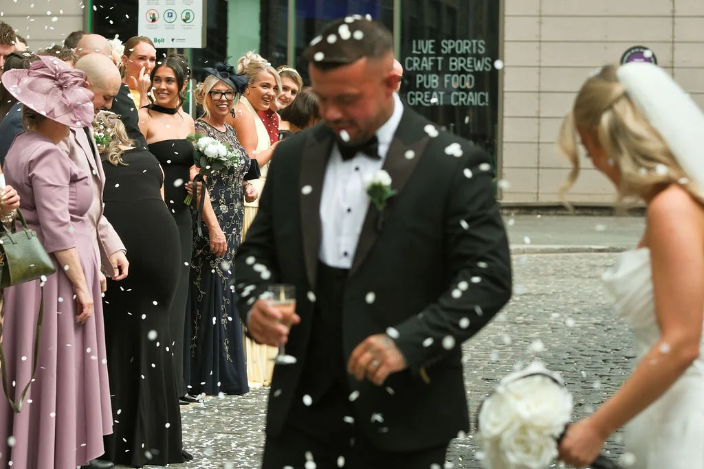 Newlyweds make their exit through a shower of white confetti as cheering guests line a cobbled city street.