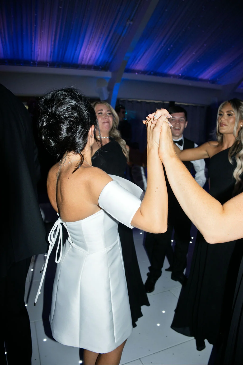 A bride in a short white off-the-shoulder dress with a lace-up back dances on a lit dance floor, holding hands with guests during the evening reception.