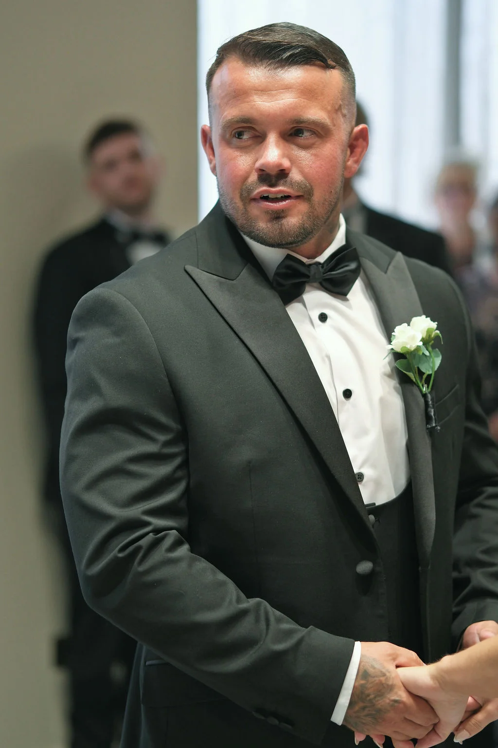 A groom wearing a classic black tuxedo and bow tie with a white rose boutonniere looks intently during the ceremony.
