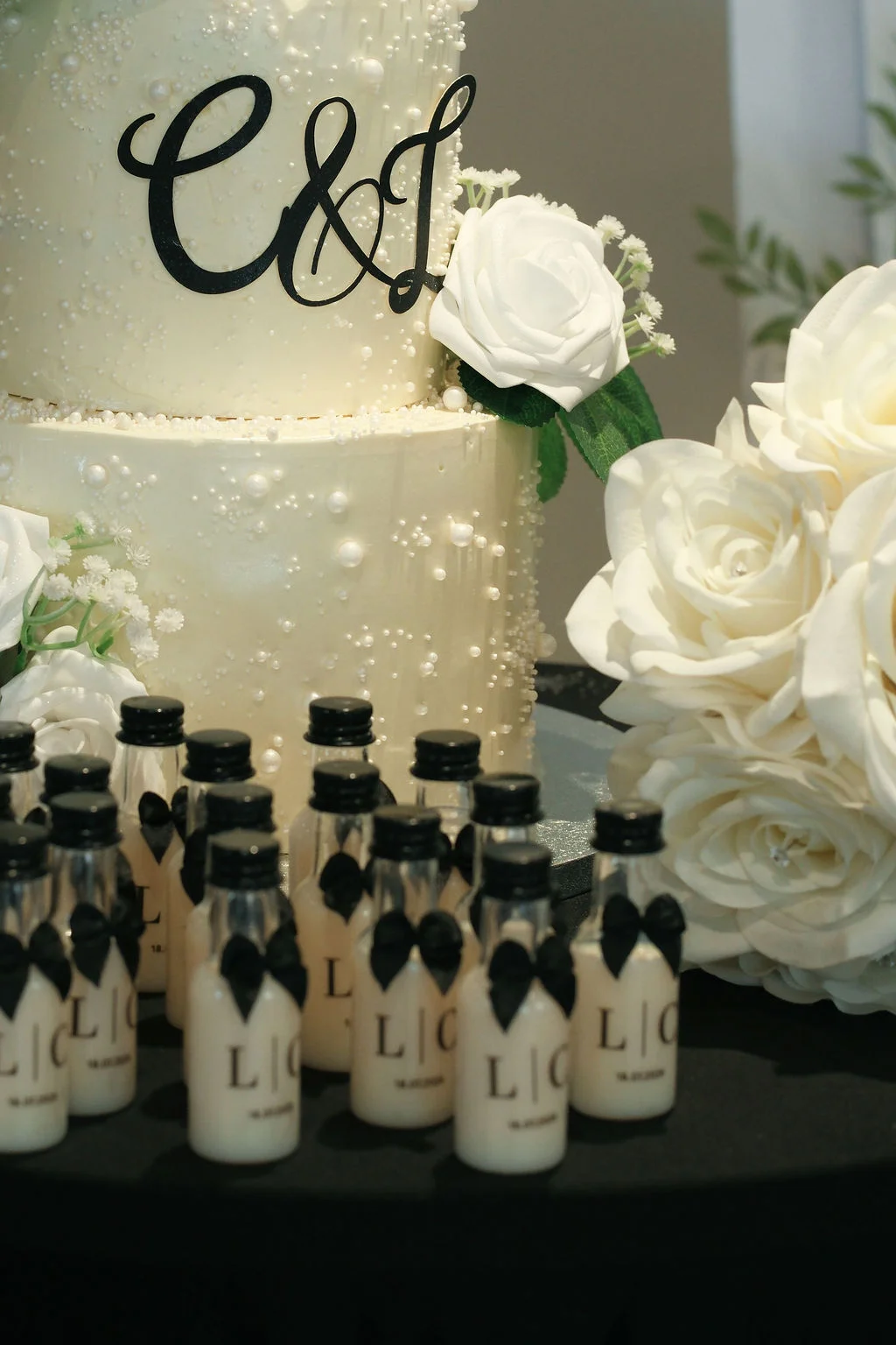 A detailed close-up shot of a white multi-tiered wedding cake adorned with edible pearls and a black custom monogram topper, displayed alongside rows of miniature favor bottles and white roses.