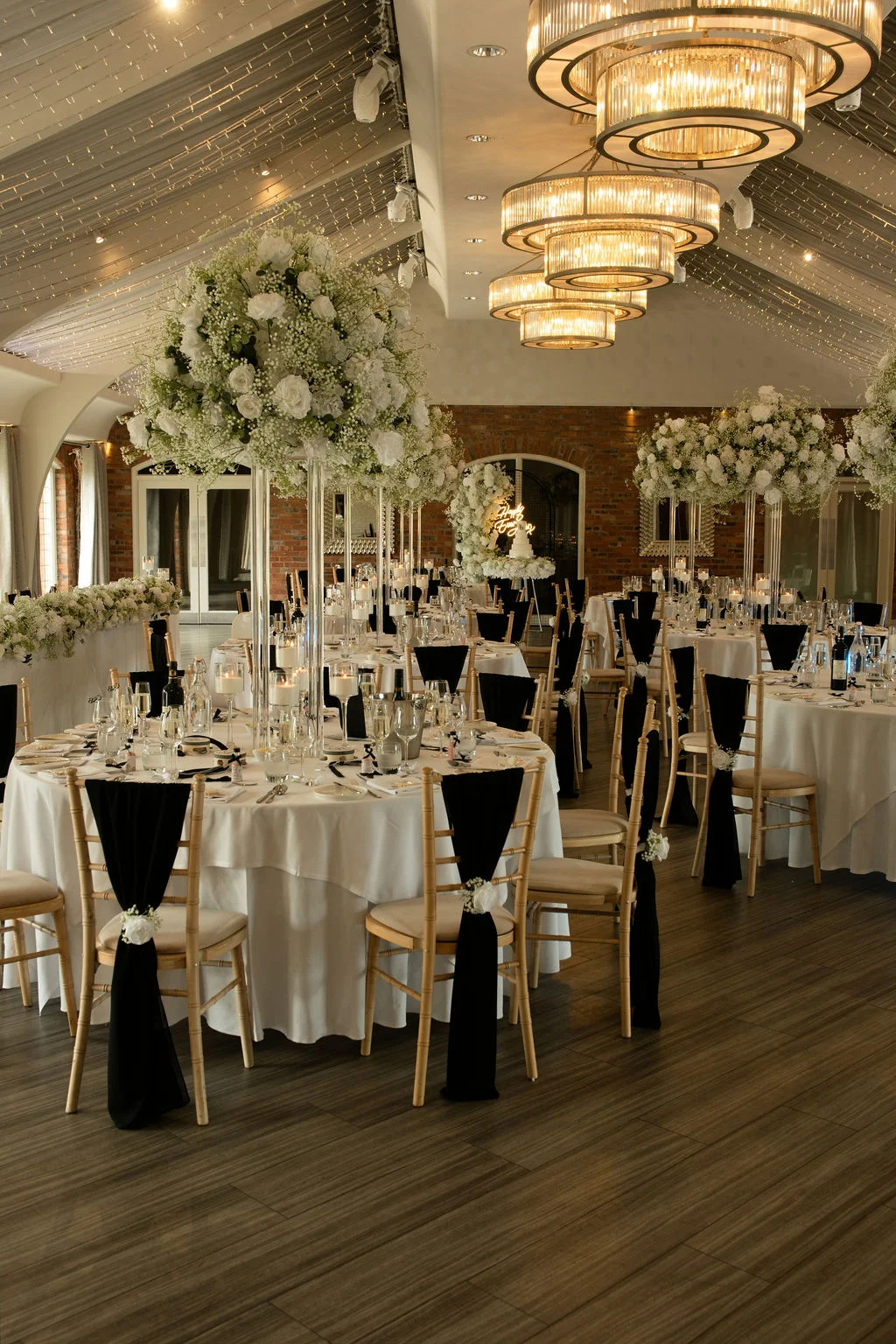 An elegant wedding reception room features round tables dressed in white linens, black chair sashes, and dramatic tall floral centrepieces beneath a sparkling fairy light canopy.