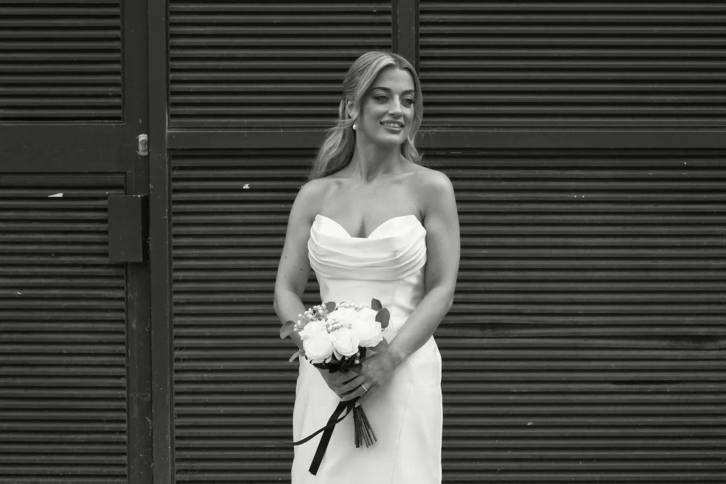 A black and white portrait of a smiling bride holding a bouquet of white roses against a slatted door background.