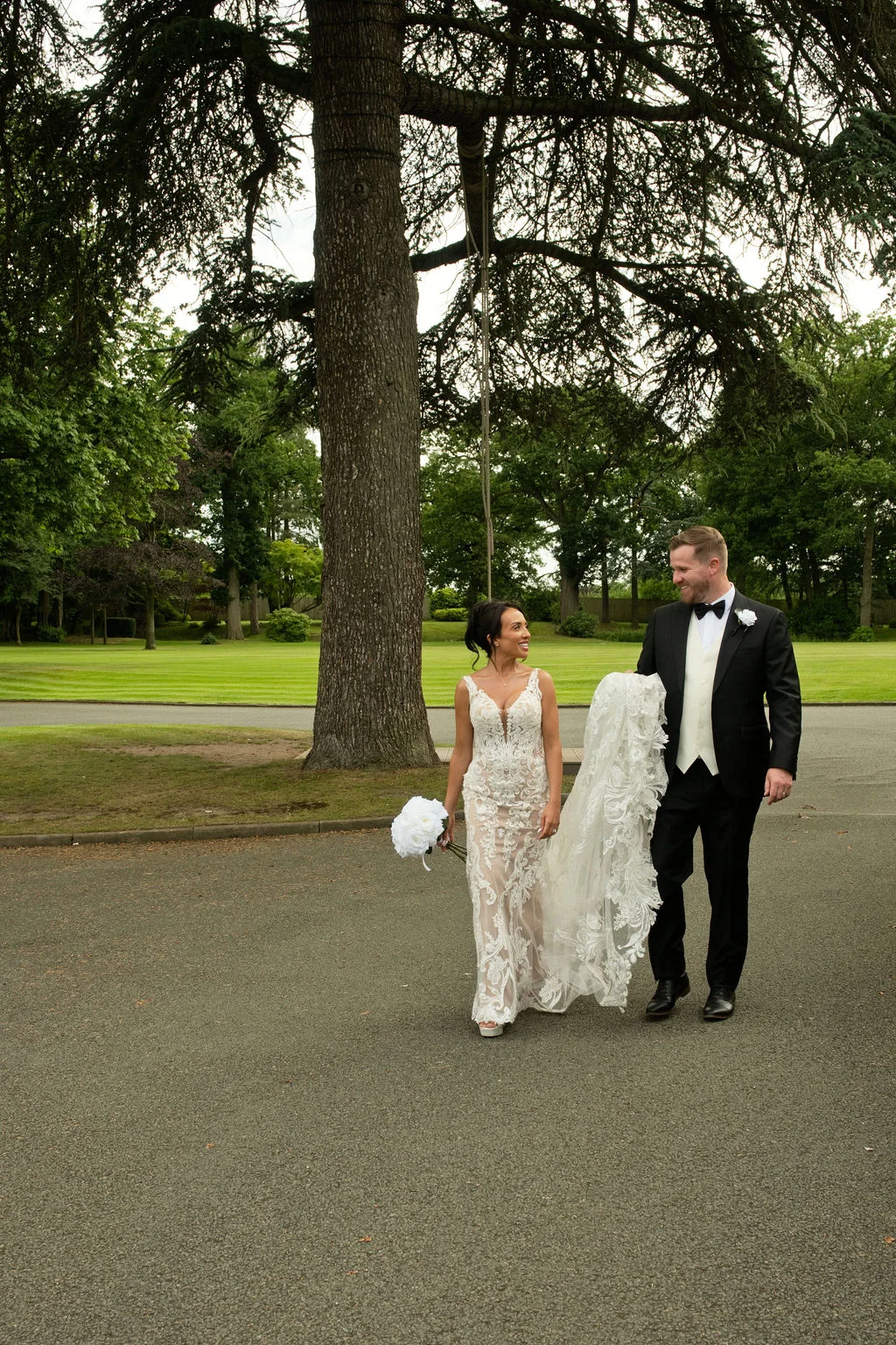 A newlywed couple smiling at each other while walking together along a paved pathway beneath a large tree on a bright summer day.
