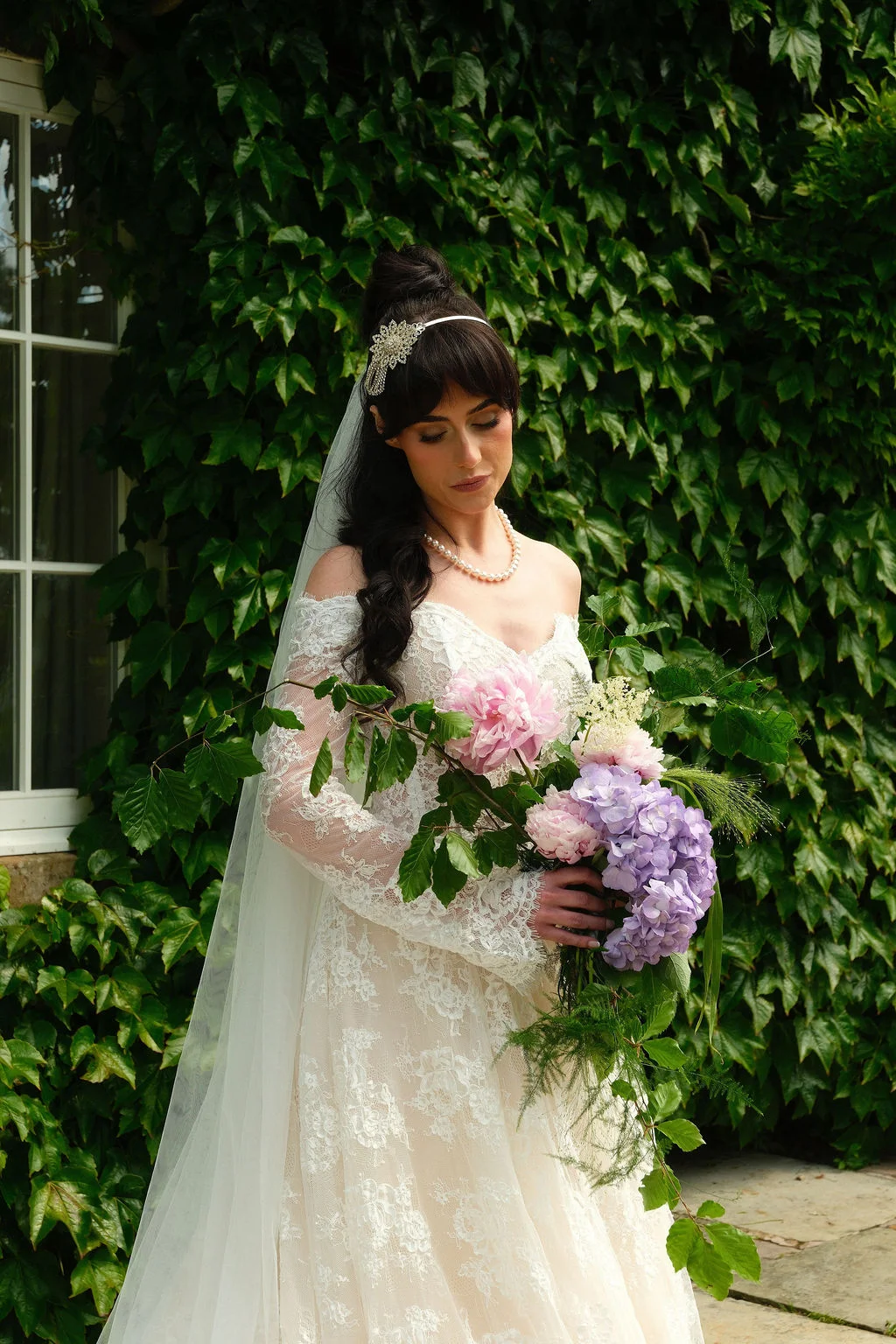 A stunning bride poses in front of an ivy-covered wall, wearing an off-the-shoulder lace gown with long sleeves, a crystal tiara, and a veil while holding a lush, cascading bouquet of pink and purple flowers.