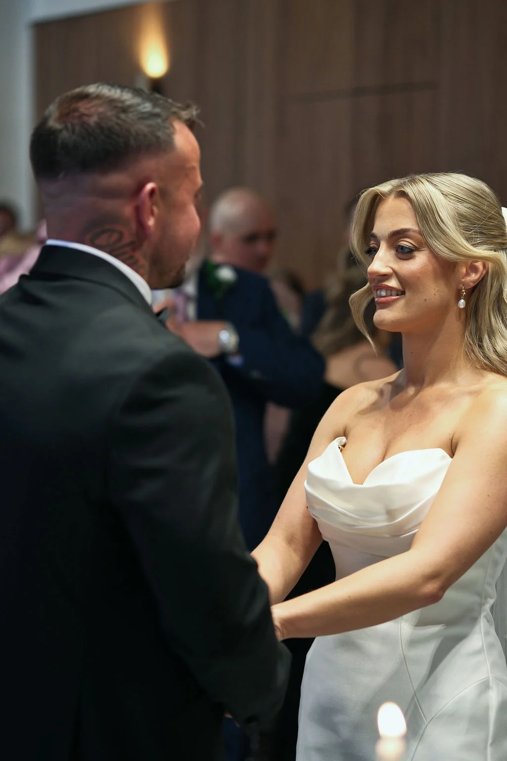 A smiling bride looks lovingly at her groom during their wedding ceremony while holding hands in front of guests.